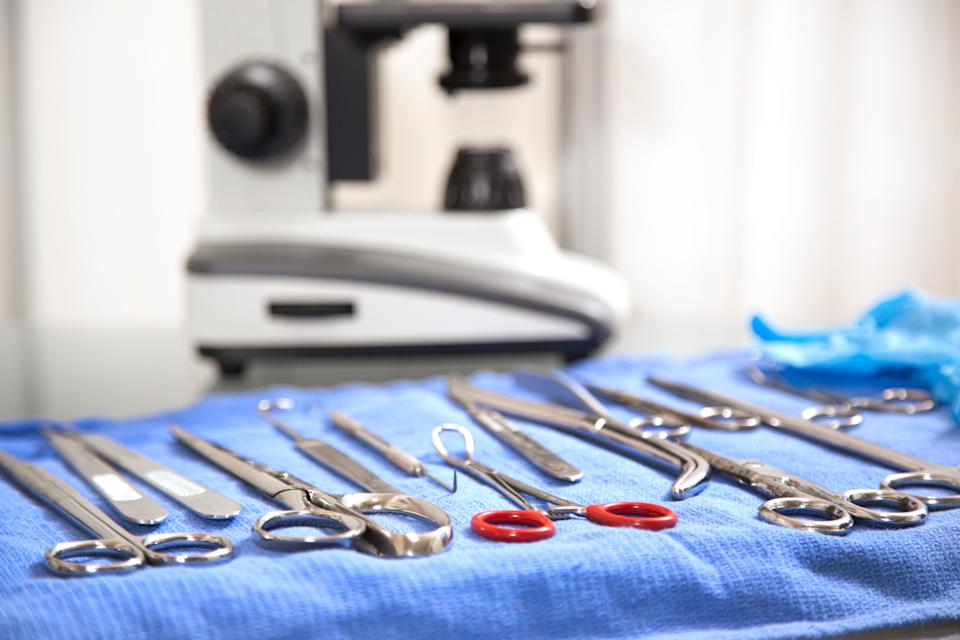 Surgical tools arranged on a blue cloth with a microscope and a pair of blue gloves in the background