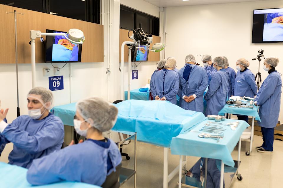 Medical professionals in a training room wearing scrubs and hairnets, focusing on a procedure shown on a monitor. Surgical tools are laid out