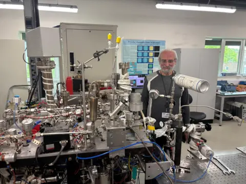BBC News A large network of tubes, metal wires, plastic cables on a table in a lab. Sasha Verchovsky stands behind the machine, which is called Finesse.