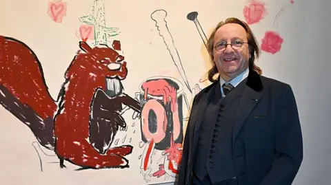 Foc Kan/WireImage/Getty Images Bill Pallot poses for a photo next to an artwork at a gallery exhibition in Paris in April. He wears a three-piece dark suit, round-rimmed spectacles and has shoulder-length hair.