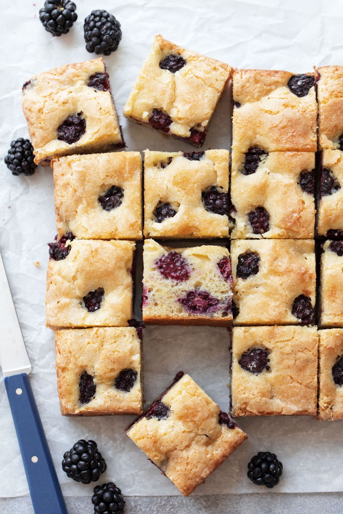 Overhead perspective, Cut squares of Blackberry Butter Cake on a piece of parchment, with a small blue knife and a few fresh blackberries scattered around.