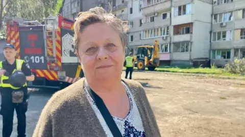 Valentina, a lady with short hair in a brown cardigan and floral shirt stood in front of the apartment building with firefighters and a truck in the background
