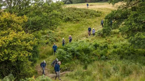 John Millard People walking down a hill in the green countryside. They are seasoned walkers of perhaps an older age and they are dressed for outdoor weather.