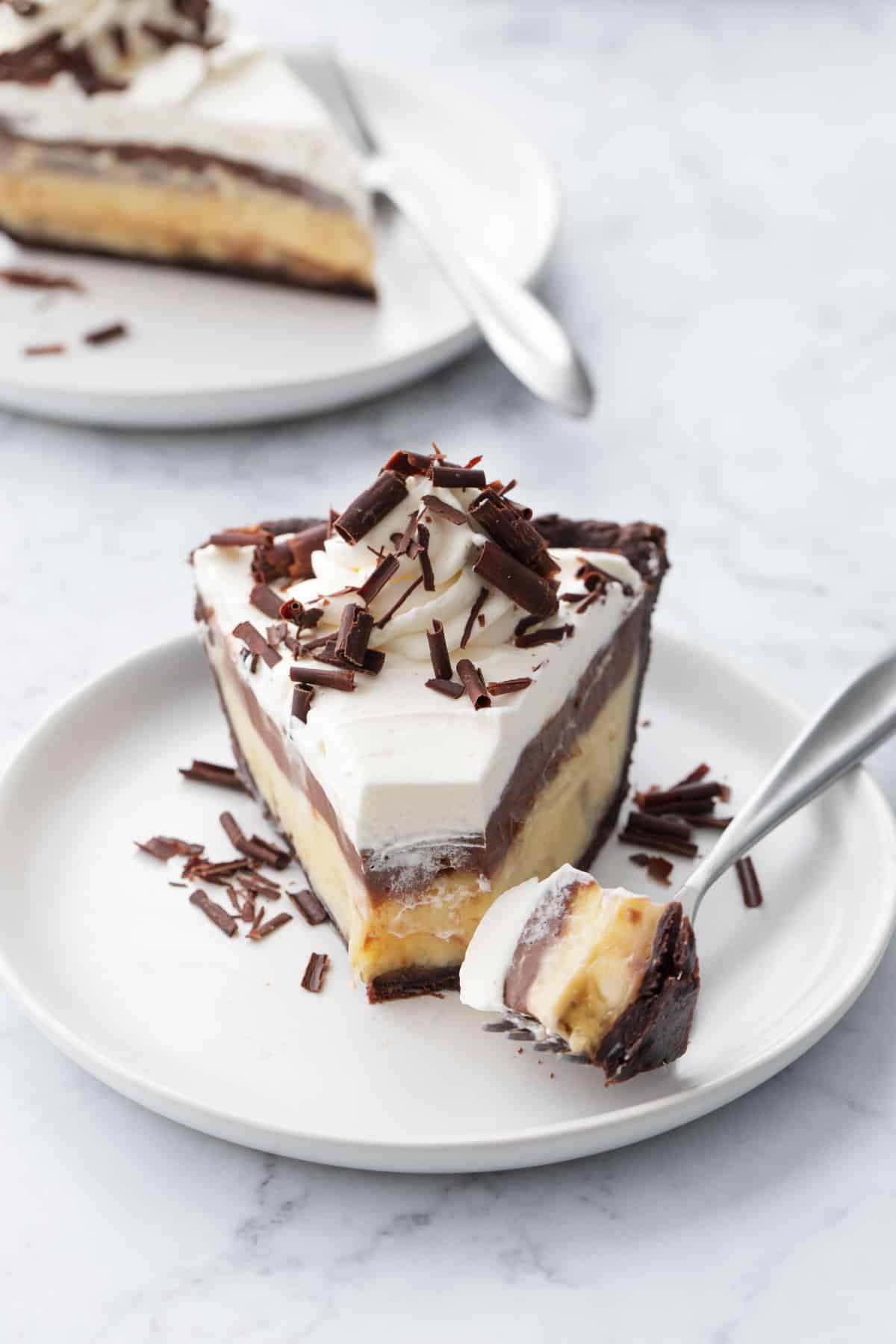 Slice of chocolate banana cream pie with a bite sitting on a fork, on a white plate on a marble background.
