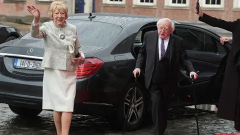 PA Media Michael D Higgins wearing a black suit with purple tie, he has two walking sticks. Sabina Higgins is wearing a cream dress and cream blazer.