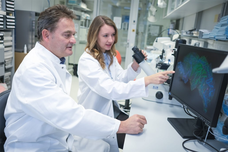 On the left, a man wearing a white coat sits at a table looking at a computer screen. Next to him, to the right, Vera Thiel wearing a white coat is sitting looking and pointing towards the same computing screen with one hand. Her other hand rests on a microscope. The computer screen has a blue and green image on it.